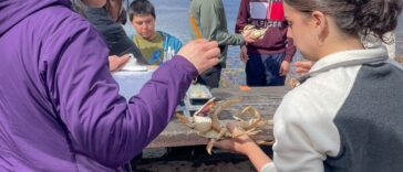 A teacher hands a crab to a high school student in an outdoor classroom at a table surrounded by students