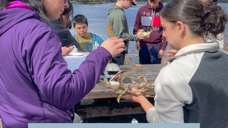 A teacher hands a crab to a high school student in an outdoor classroom at a table surrounded by students