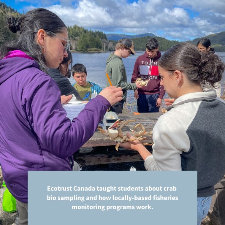 A teacher hands a crab to a high school student in an outdoor classroom at a table surrounded by students