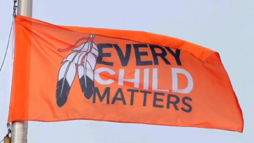 An orange flag with the text "Every Child Matters" and two black and white feathers flies against the backdrop of a blue sky